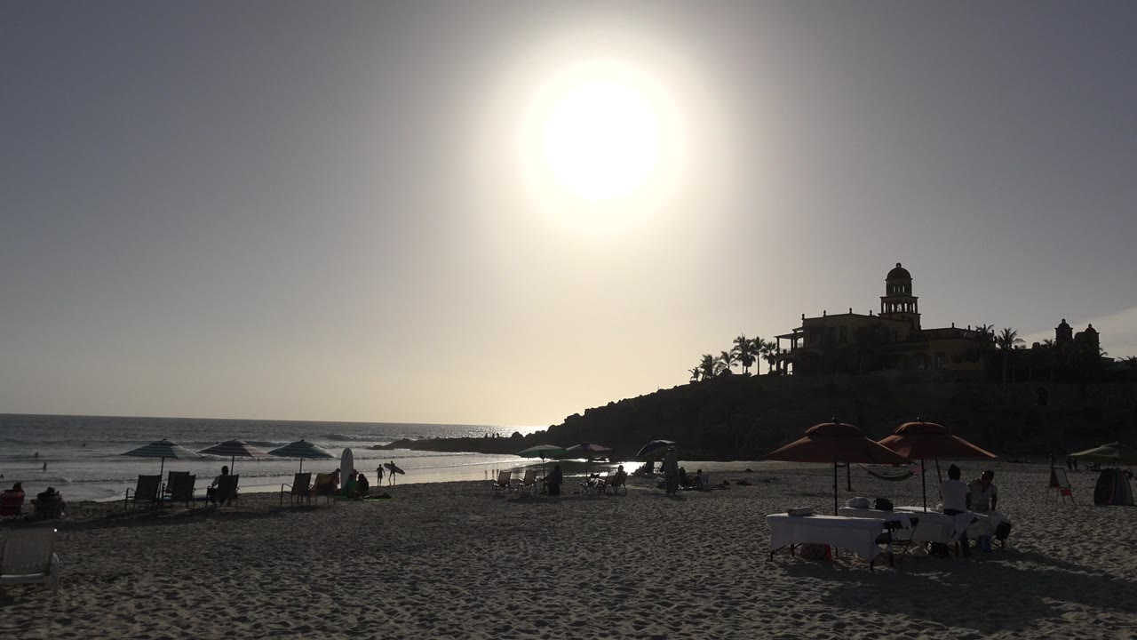 At 0:01 in Los Cabos, Mexico, the sun remains high above the ocean while parasols and chairs sit on the sand below a dark hillside with old buildings and palms.