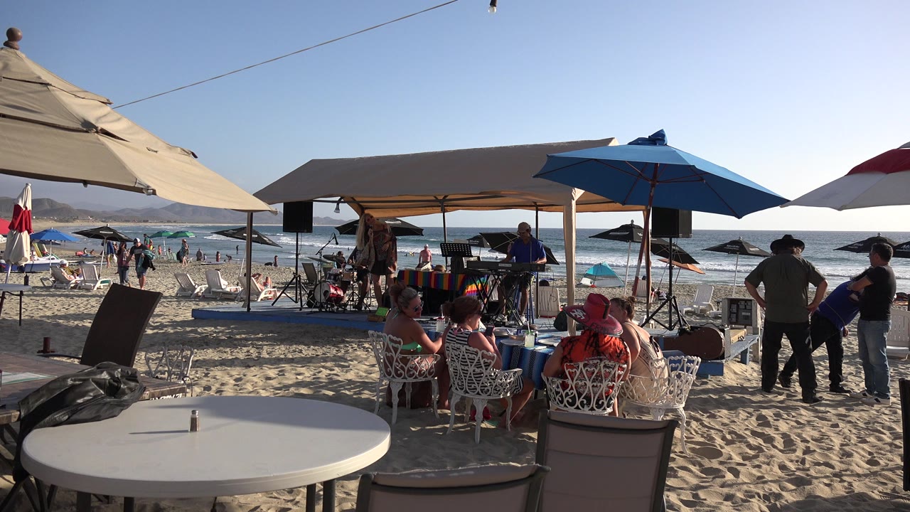 Beach umbrellas, seated adults, and ocean waves behind a sandy gathering.
