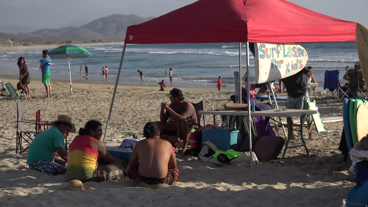 At 0:01 in Los Cabos, Mexico, the red tent fills the upper frame as surfboards and beach equipment sit near seated adults, with small groups visible along the waterline.