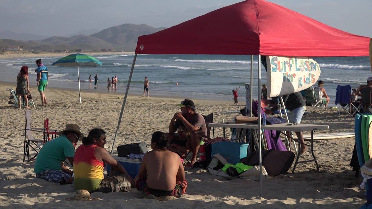 Beachgoers sitting under a red canopy with ocean and mountains behind them.