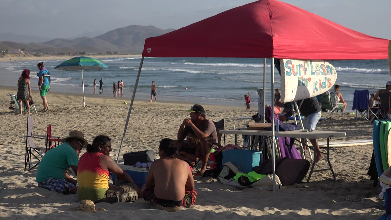 At 0:01 in Los Cabos, Mexico, the red canopy shades a seated group in the foreground as a green umbrella, shoreline walkers, and breaking waves remain visible behind them.