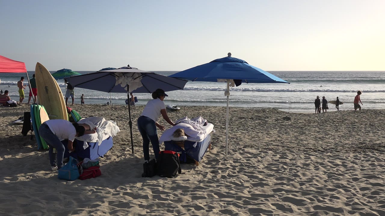 At 0:00 in Los Cabos, a beach massage table sits under blue and gray umbrellas beside upright surfboards, with people visible near the waves.