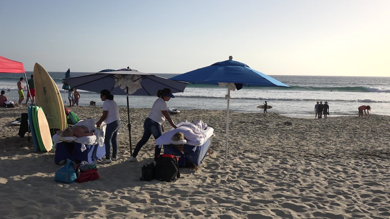 Beach umbrellas, surfboards, towels, and people near the ocean at sunset.