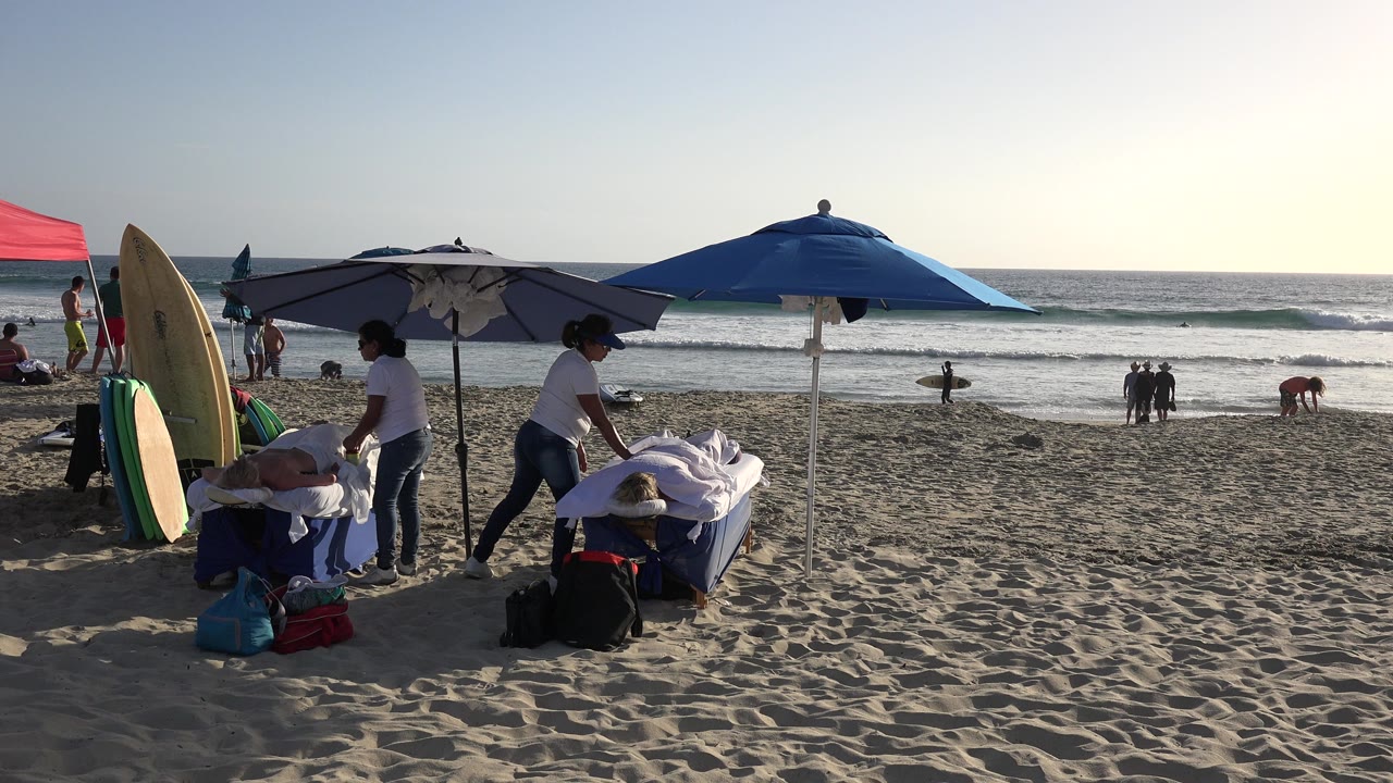 Los Cabos beach frame at 0:01 with a person in a white shirt near a towel-covered lounge area, upright surfboards on the left, and several people by the waterline.