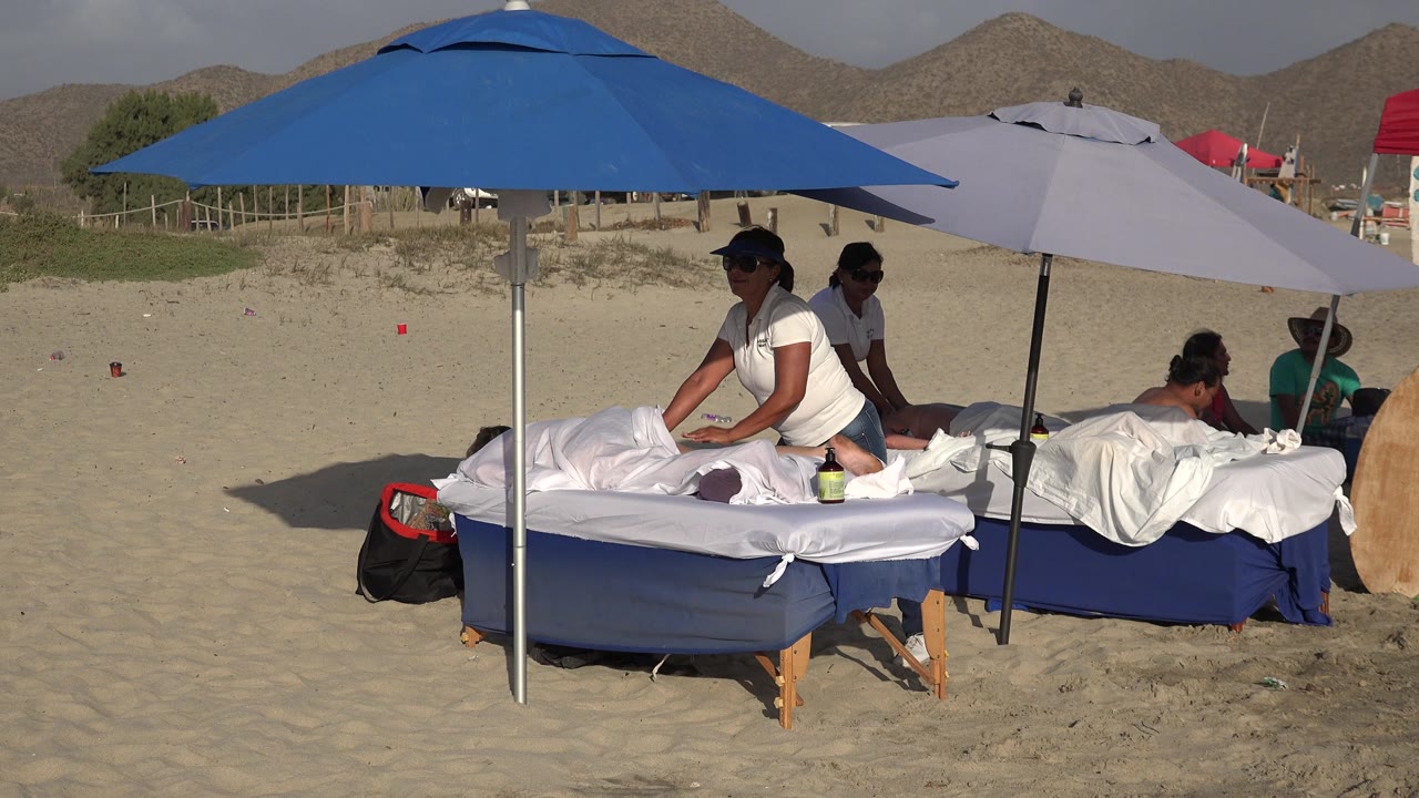 Los Cabos beach scene with a therapist leaning over the foreground massage table beneath a blue umbrella while other people sit or recline near a pale umbrella.
