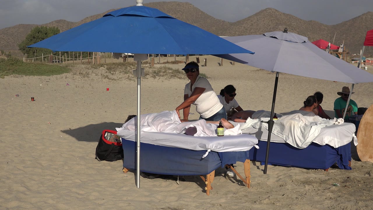 Opening frame of a Los Cabos beach massage setup with a blue umbrella over a towel-covered table, a pale umbrella beside it, attendants and clients visible, and low hills behind the sand.