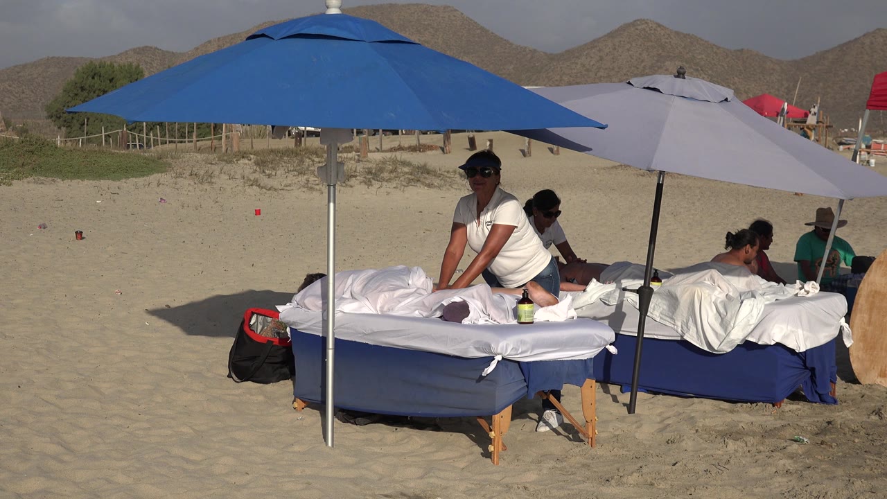 Beach massage scene with attendants, clients, umbrellas, and sandy background.