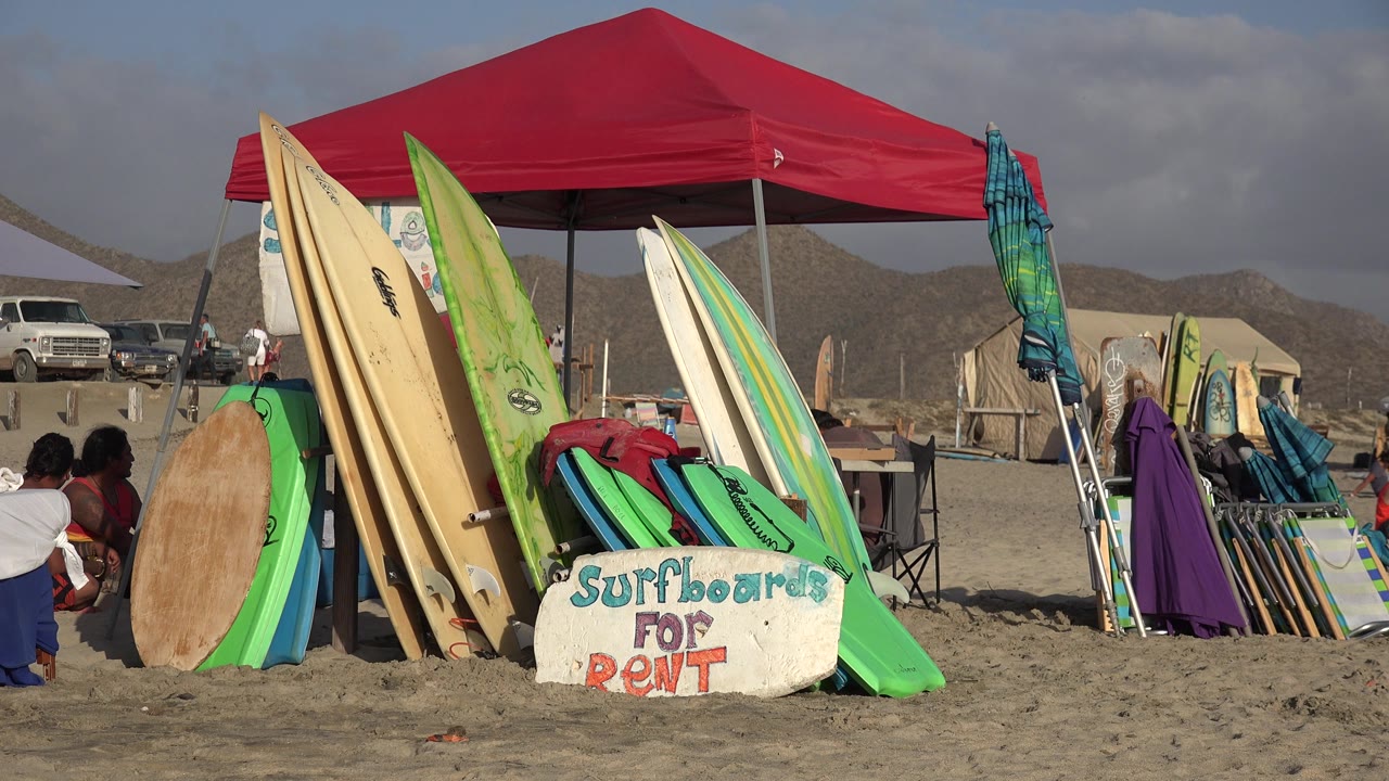At 0:01 in Los Cabos, Mexico, the rental stand remains centered with tall surfboards, green boogie boards, a red canopy, and seated or crouching people near the left side of the frame.