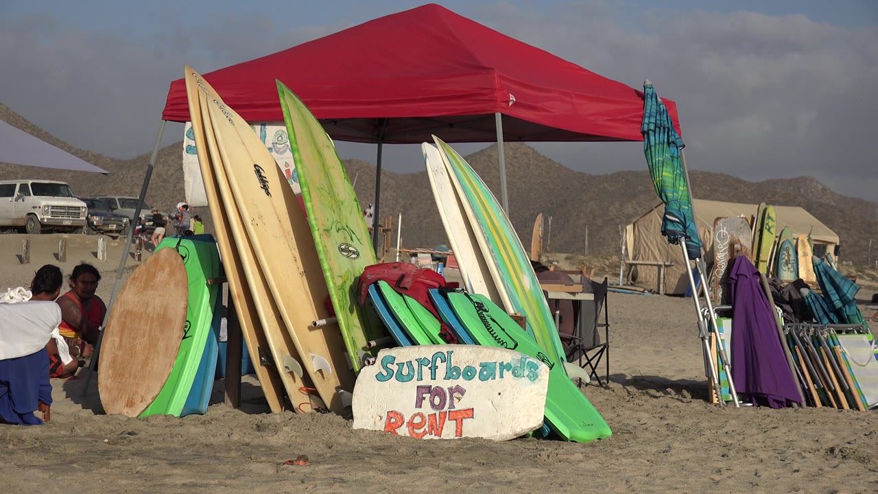 At 0:00 in Los Cabos, Mexico, a red canopy tent stands behind upright surfboards and a hand-painted surfboards for rent sign, with seated people partly visible at the left edge.