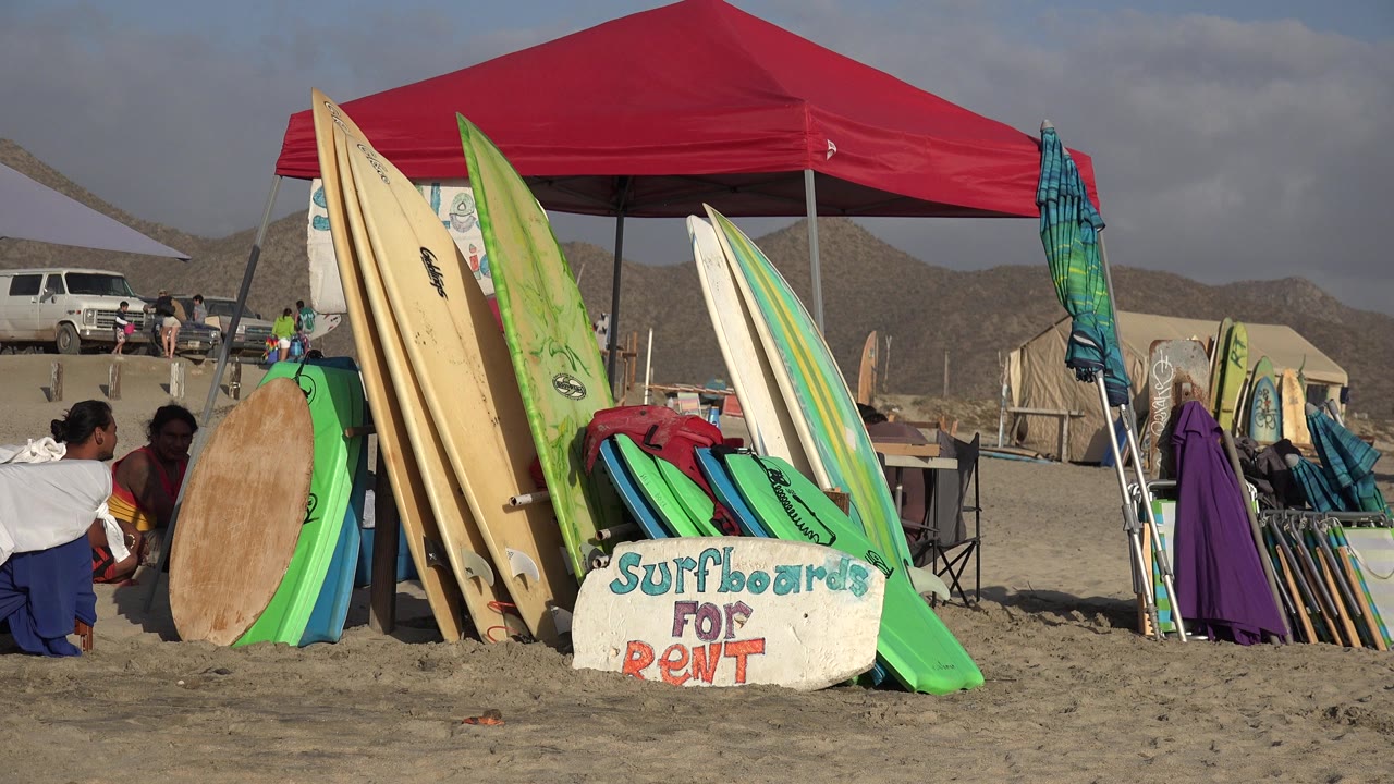 Surfboards stacked beside a red beach canopy with a rental sign in the sand.