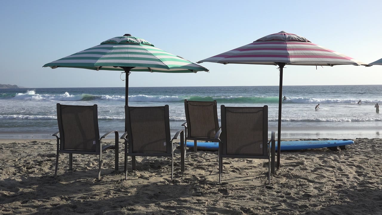 At 0:01, the same Los Cabos beach setup holds steady as a wave line advances behind the chairs and distant bathers remain small in the background.