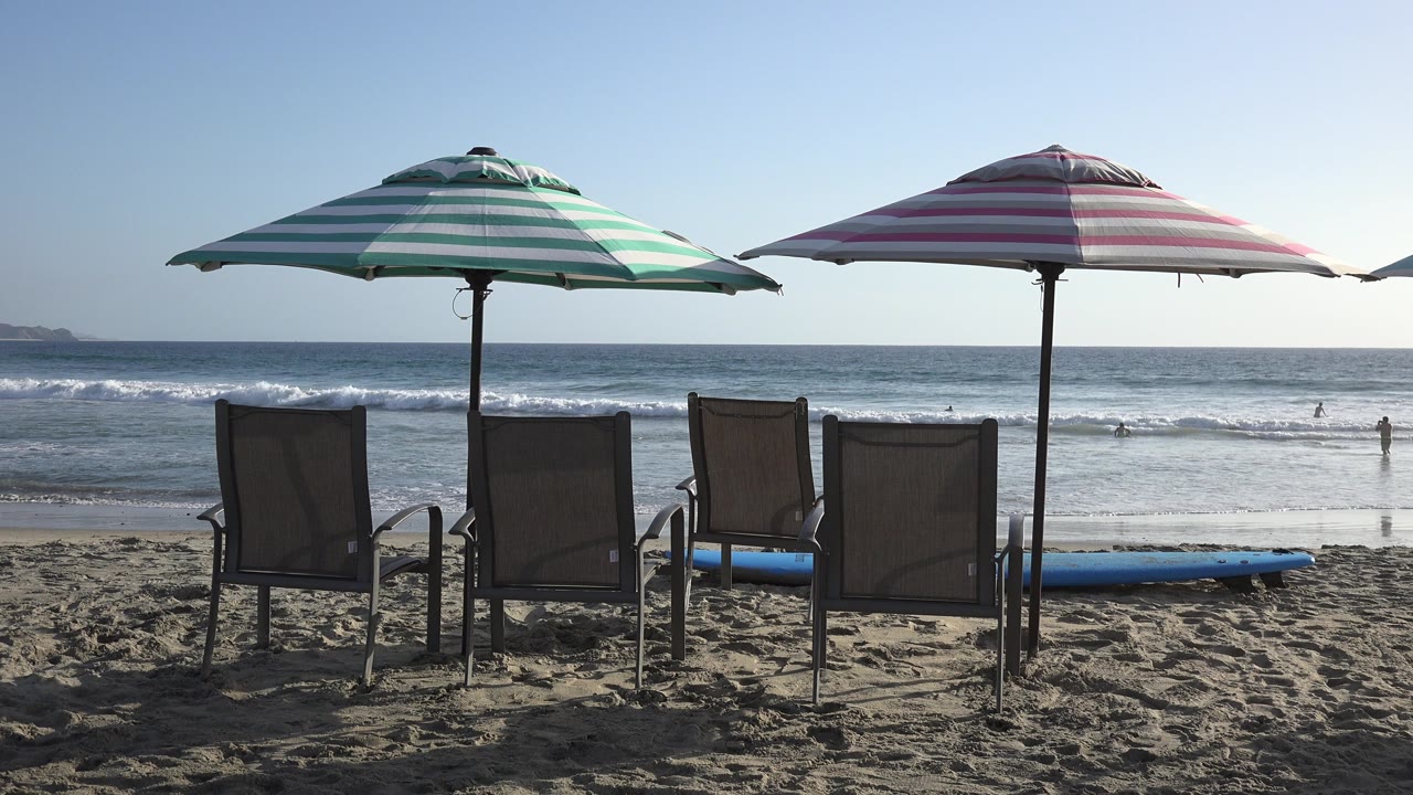Los Cabos beach view showing four lounge chairs beneath green and pink striped umbrellas, with white surf bands moving across the ocean background.