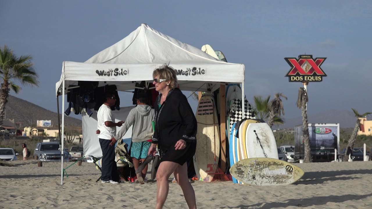 Woman walking past a surfboard vendor tent on a sandy beach.
