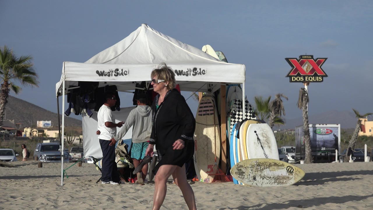 At 0:01 in Los Cabos, Mexico, several people stand near upright surfboards under a white canopy while a woman continues walking across the sandy foreground.