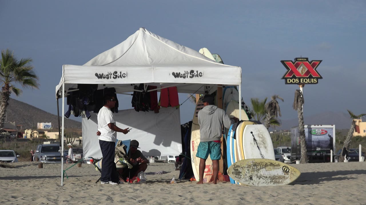 Los Cabos beach scene with one person standing outside the white pavilion, others seated in shade, surfboards at the entrance, and the Dos Equis sign behind the tent.