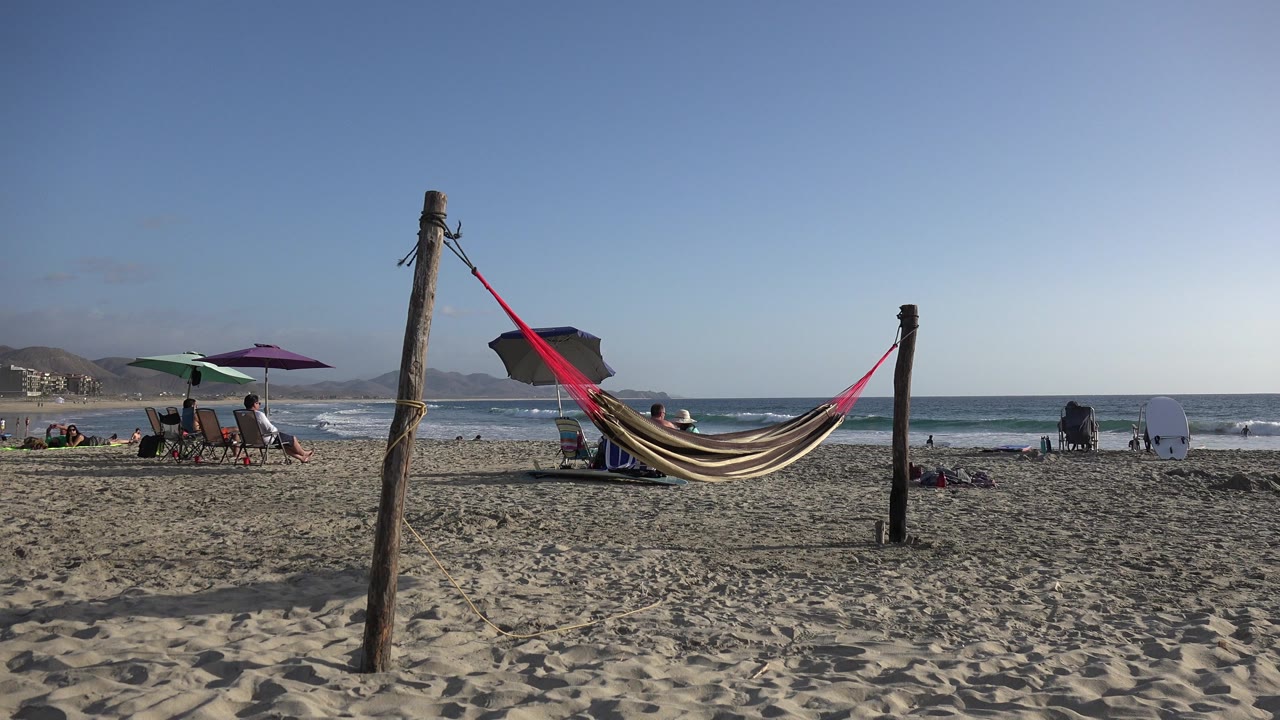 The hammock remains centered in the Los Cabos beach scene as small groups sit under umbrellas near the surf and white waves roll across the background.