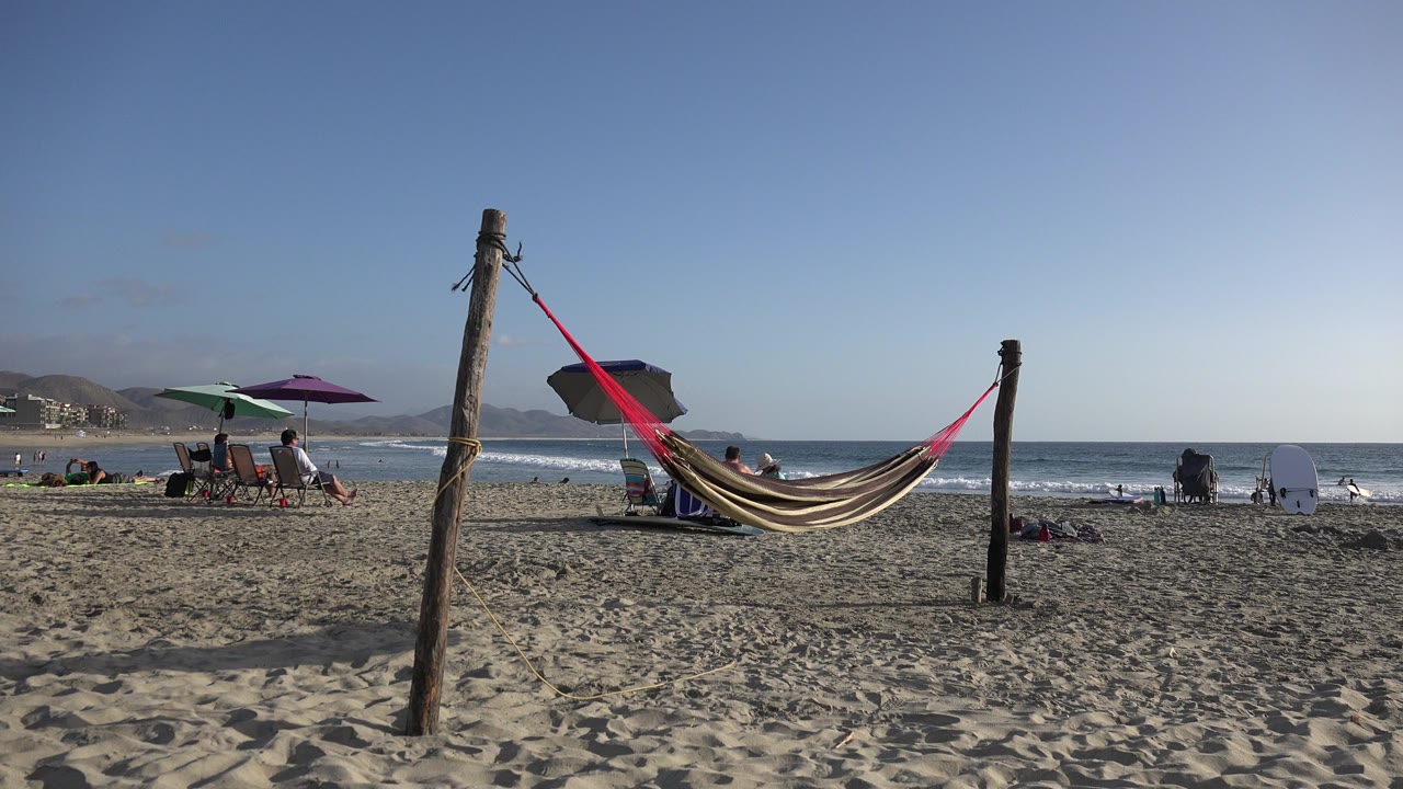 Hammock between wooden posts on a sandy beach with ocean and umbrellas behind it.