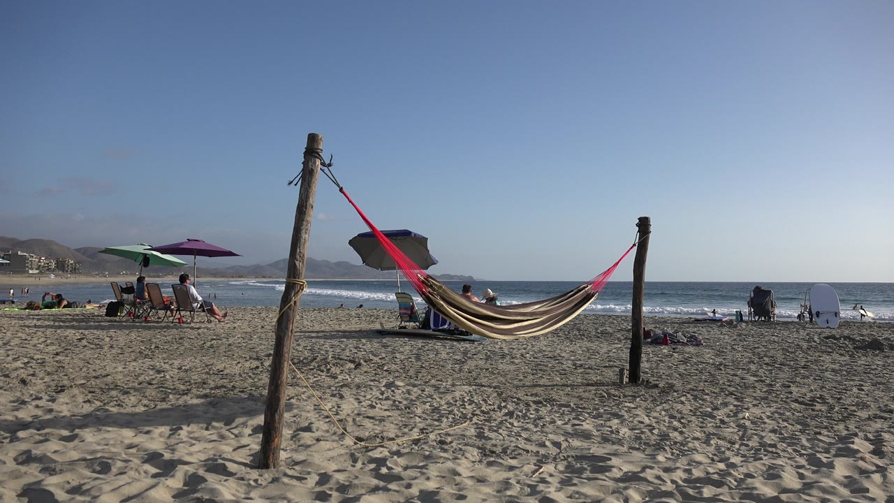 The hammock remains centered in the sandy foreground as people sit under purple and green umbrellas near the Los Cabos shoreline.