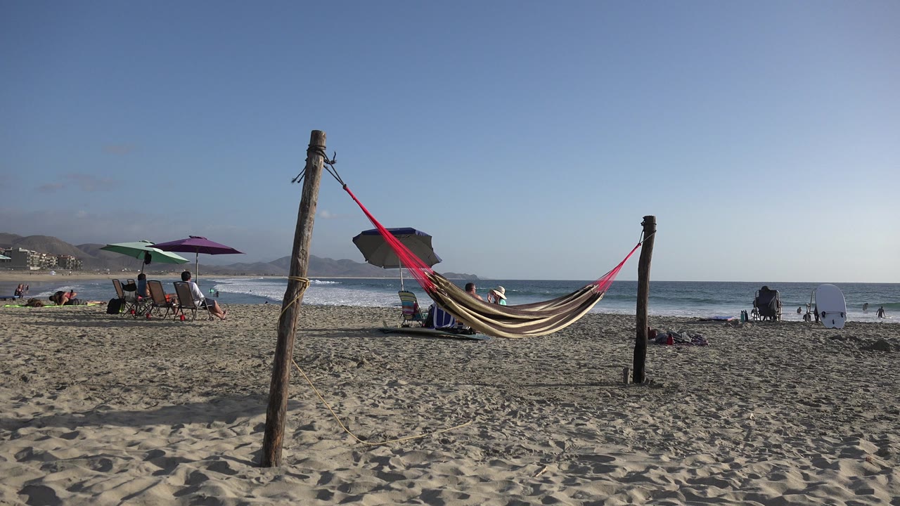 Hammock between wooden posts on a sandy beach with umbrellas and ocean behind it.