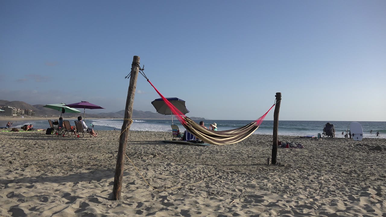 The Los Cabos beach view holds on the hammock and posts as beach chairs and umbrellas sit near the shoreline with a few people visible under the blue sky.