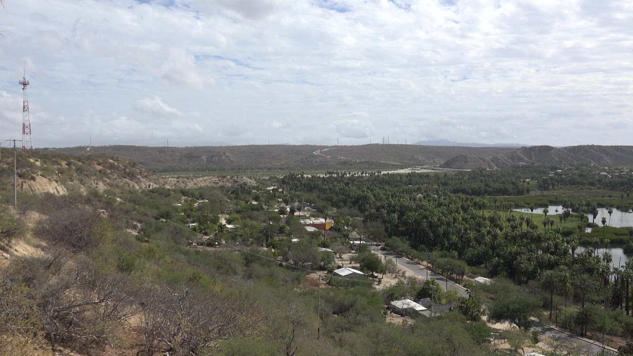 Second frame shows the Los Cabos hillside overlook with a clearer central valley of palms, small buildings, pale roads, reflective ponds, distant hills, and no visible people.