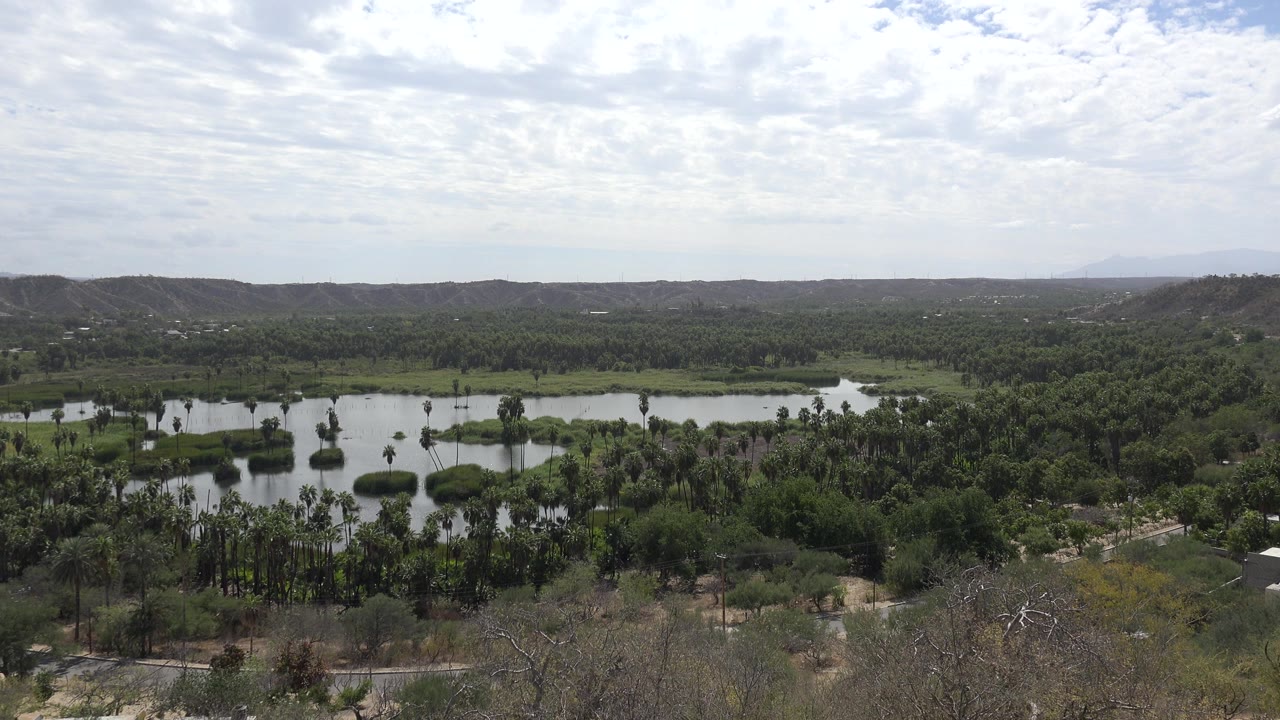Opening wide view of a green Los Cabos landscape with small bodies of water, dense trees, dry hills, a bright clouded sky, and no visible people.