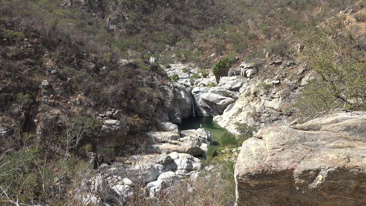 Los Cabos, Mexico creek below pale rocks and dry hillside brush, with a large boulder at right and no visible people.