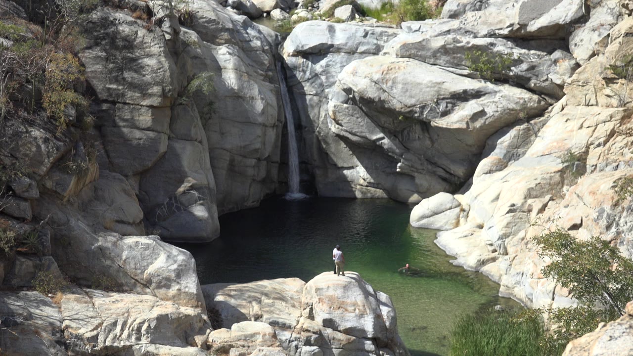 In Los Cabos, Mexico, a person stands on a rounded rock above green water while a small waterfall drops between pale boulders in the background.