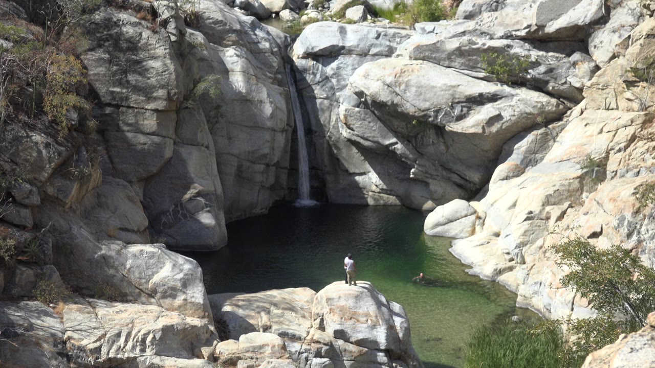 A slightly later Los Cabos frame keeps the person on the lower rock in view, with the swimmer farther across the green pool and the waterfall shaded by the cliff.