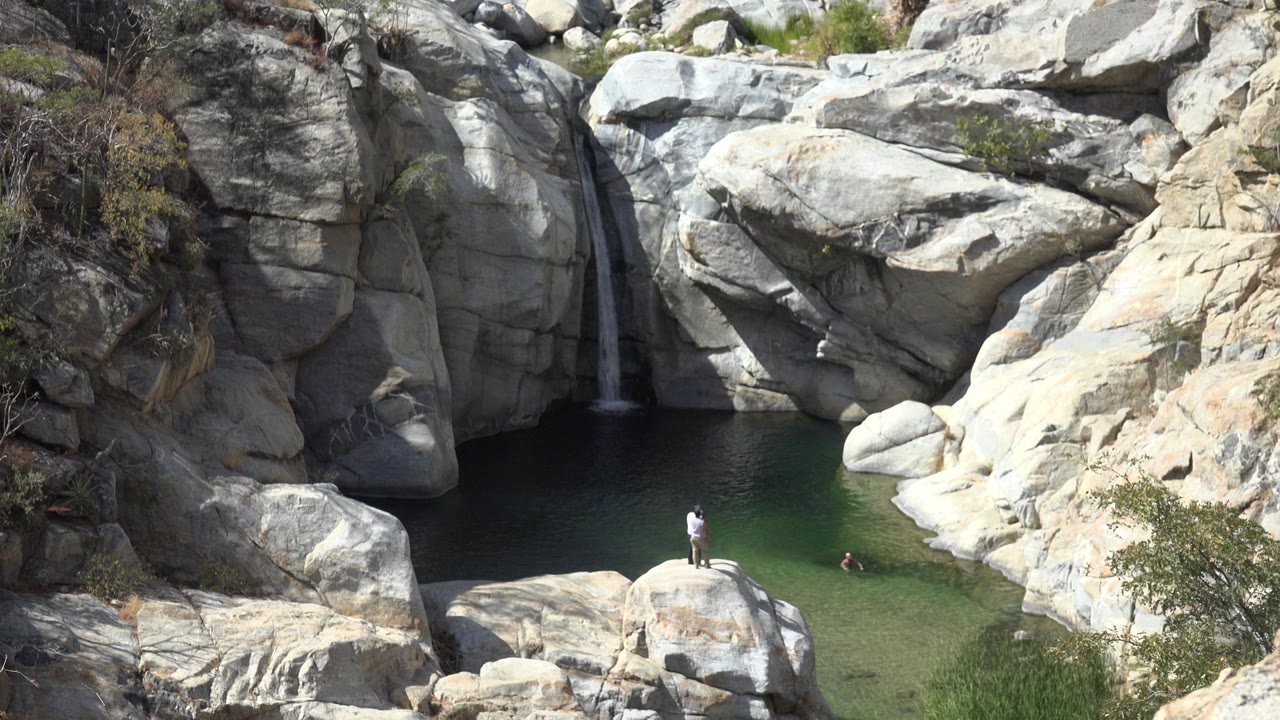 Los Cabos, Mexico rocky pool with a narrow waterfall at left, a person standing on a boulder, and swimmers visible in the green water.