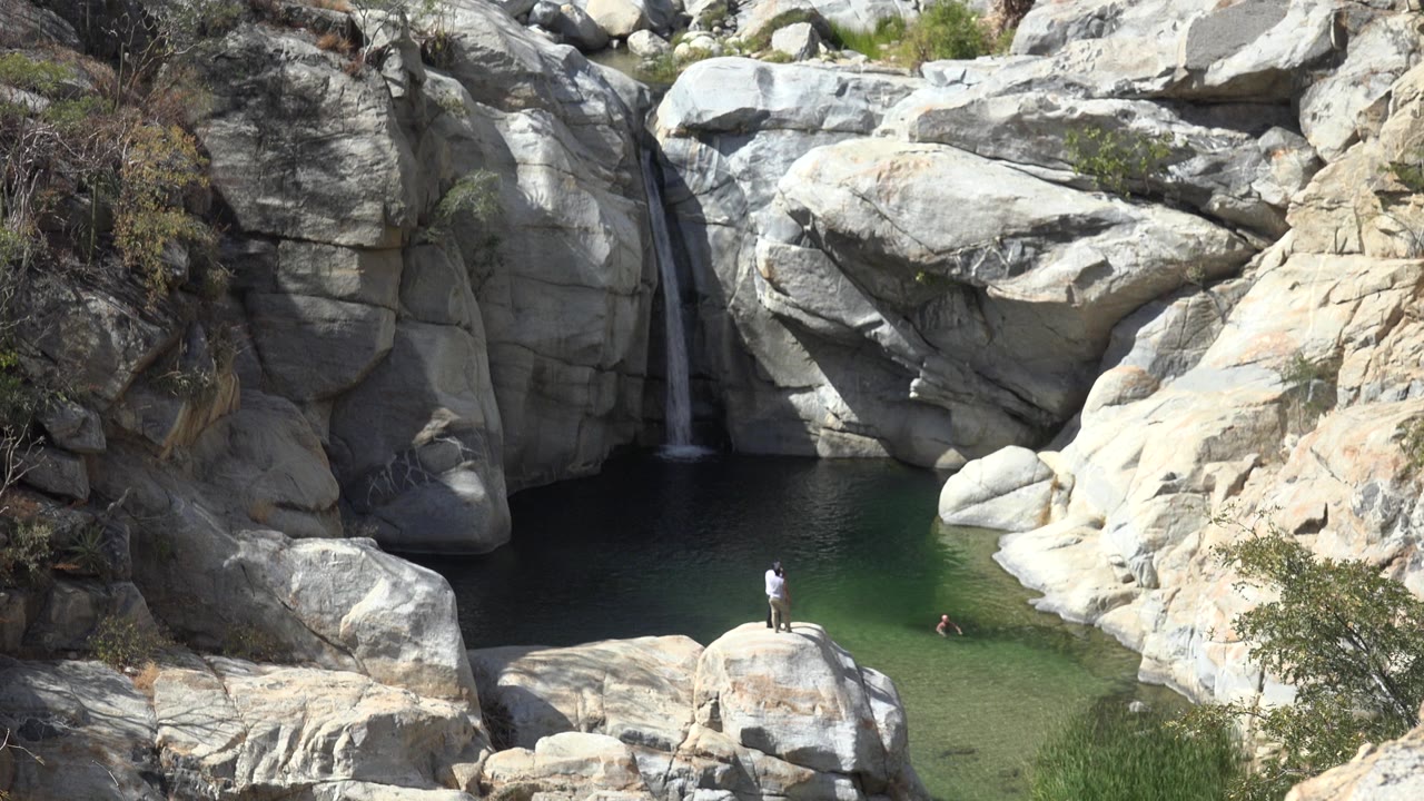 Los Cabos, Mexico waterfall scene with the standing person still on the foreground boulder and a swimmer visible near the right side of the pool.