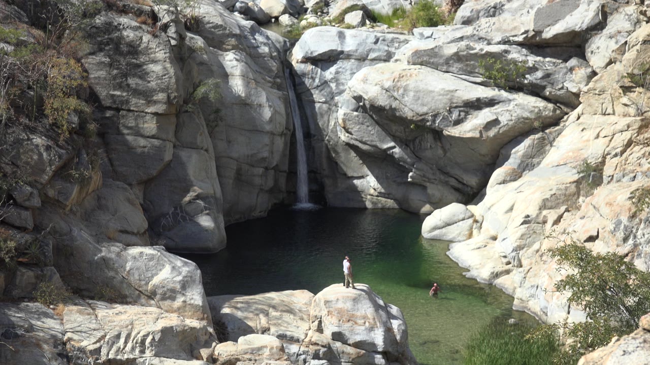Los Cabos rocky pool with pale boulders, a narrow waterfall at left, and small people visible on a rock and in the water at the start of the clip.