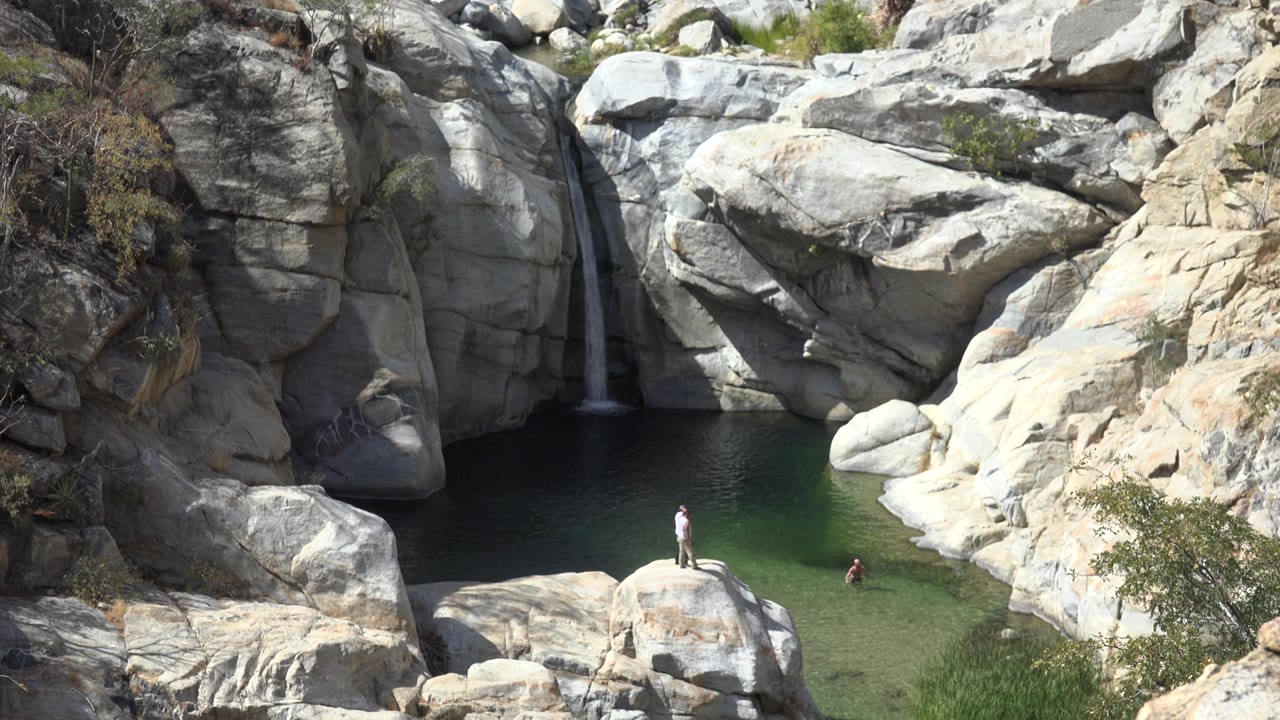 Los Cabos waterfall drops into a dark green pool between pale rock faces, with small people visible near the lower right side of the water.