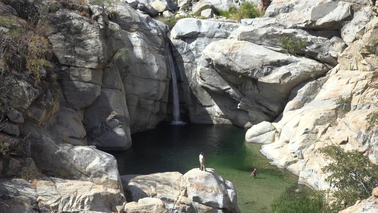 Wide view of boulders, waterfall, and green pool with small figures.