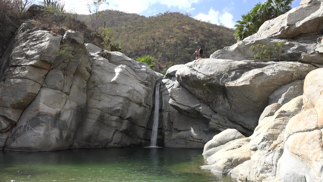 A Los Cabos waterfall drops between pale boulders into green water, with dry hills and a tiny distant person on the upper rocks.