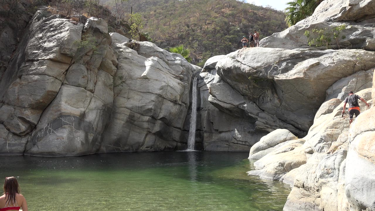 Rocky pool with a narrow waterfall and people on the rocks.