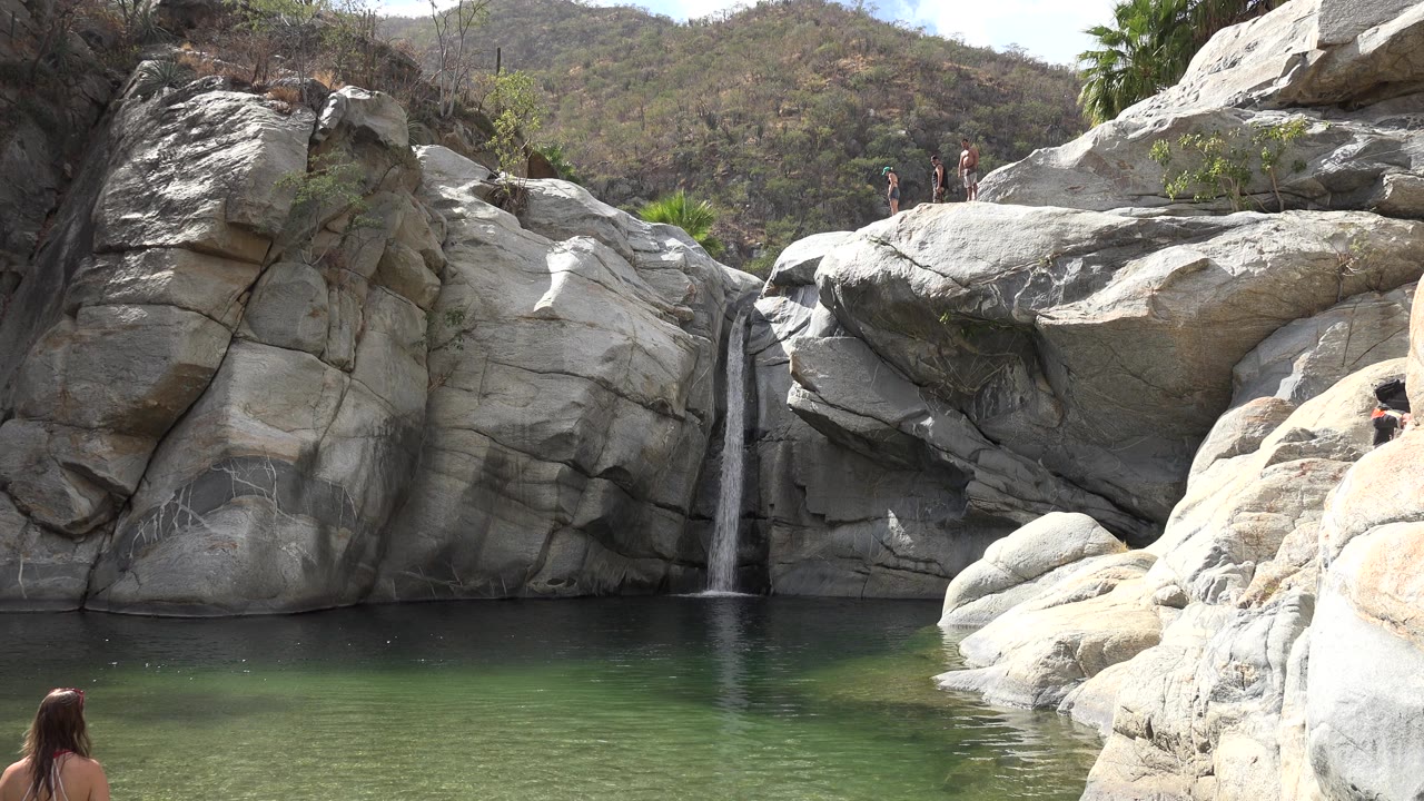 At the start of the Los Cabos clip, a narrow waterfall drops between pale rocks into green water while small people stand on upper ledges and a woman is partly visible at lower left.