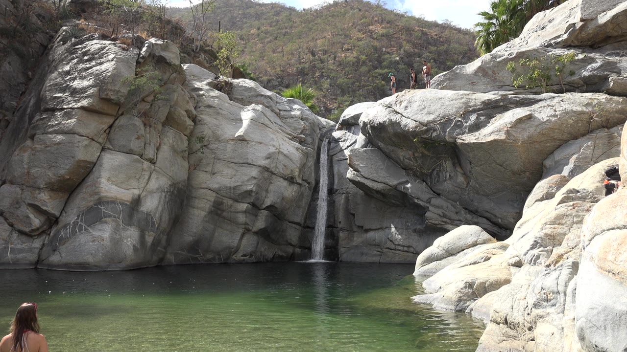 One second in, the Los Cabos waterfall remains centered between pale rock walls, with green water below, distant hills above, and several small visitors standing along the upper rock shelf.