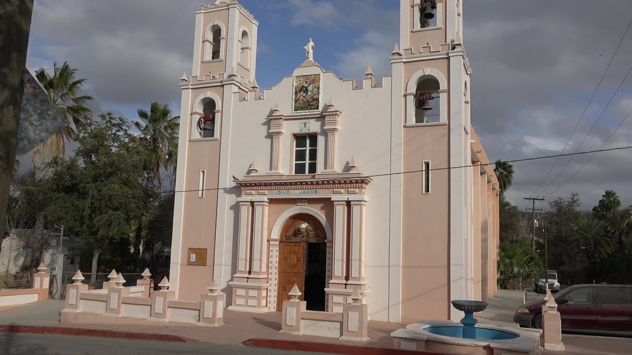 Pale church facade with bell towers, fountain, cloudy sky, trees, and street.