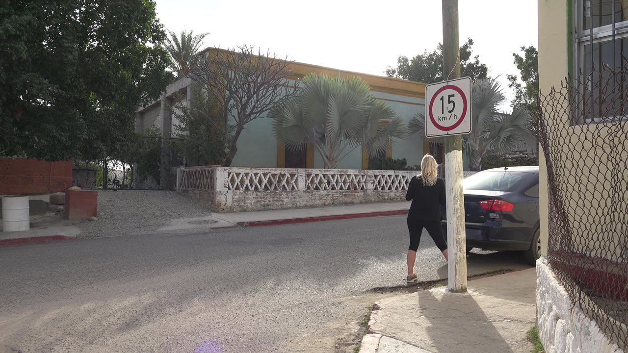 Blonde woman in black stands near a utility pole with a 15 km/h sign on a quiet Los Cabos residential street, with a parked car and houses behind her.