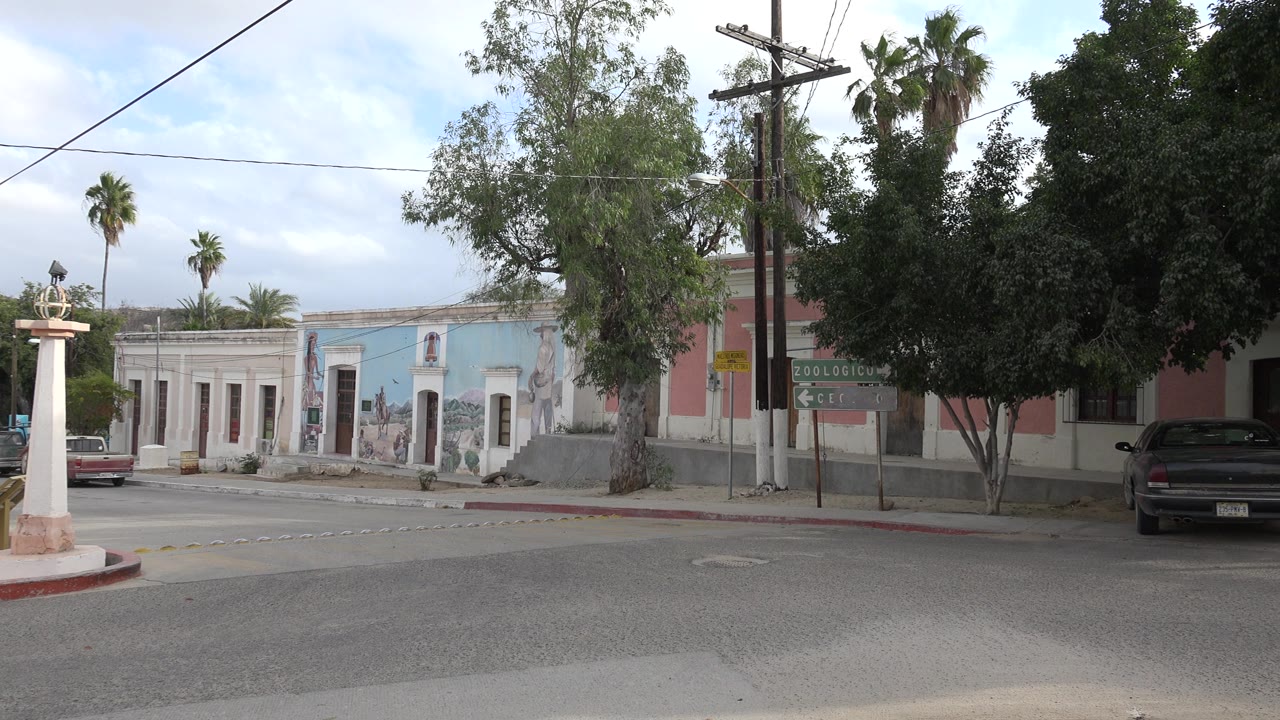 Los Cabos street corner with a white column near the curb, a blue mural building across the road, palm trees in the background, scattered cars, cloudy sky, and no visible people.