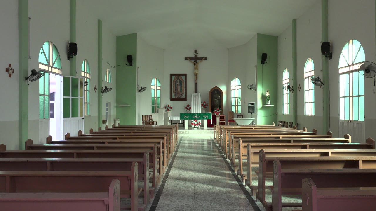 At 0:01, the Los Cabos church view remains centered on the aisle, with pew backs in the foreground, bright side windows, wall-mounted fans, the altar, a crucifix, and no visible people.