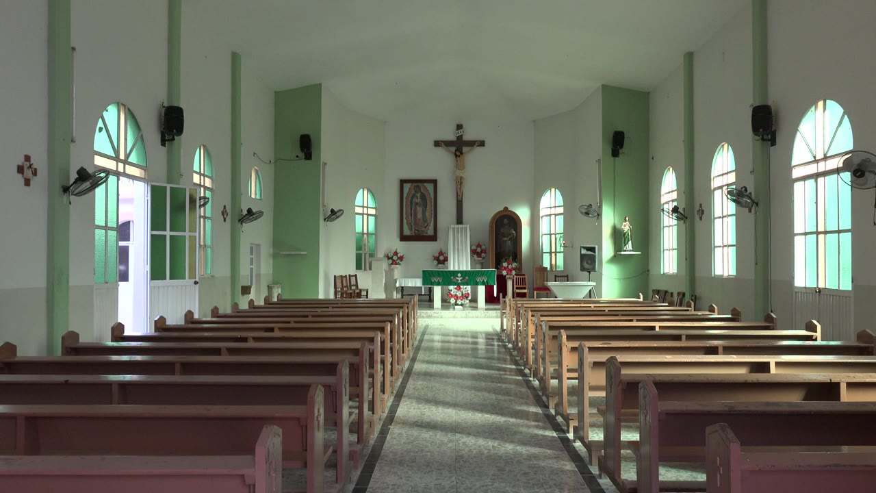 Interior frame from the Los Cabos church looking past rows of pews toward the altar, crucifix, side windows, and no visible people.