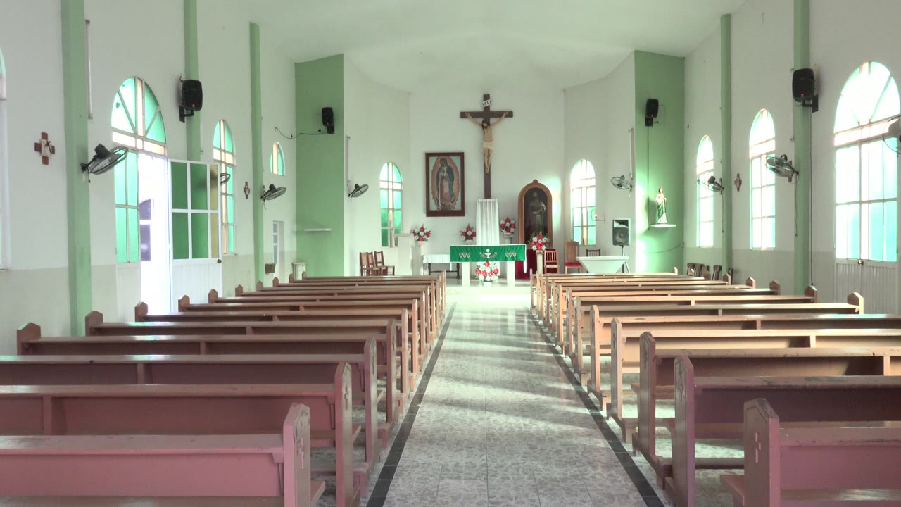 Rear view inside a Los Cabos Catholic church shows rows of wooden pews leading to a green-draped altar, crucifix, arched windows, and no visible people.