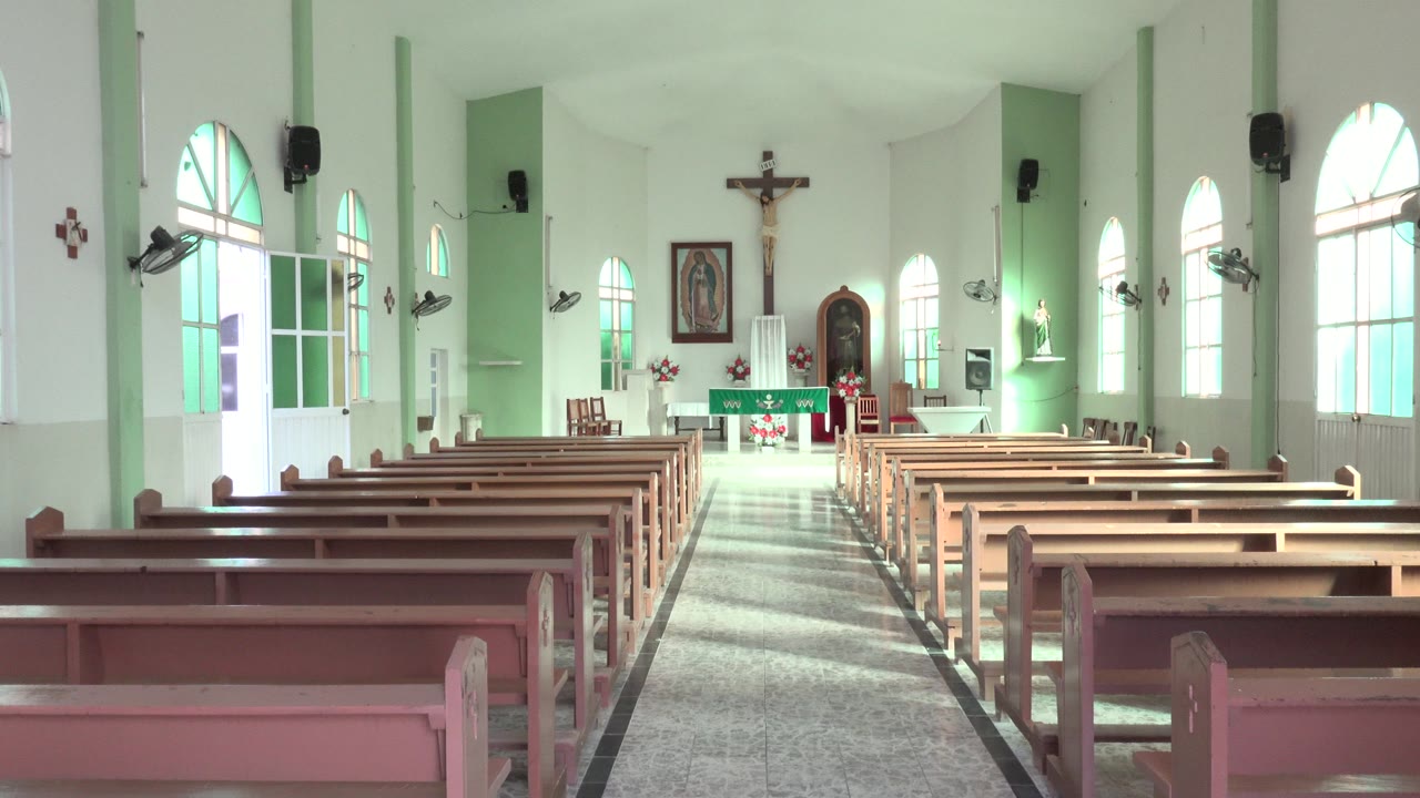 Empty nave with arched windows, long pew rows, altar flowers, and wall-mounted fixtures.