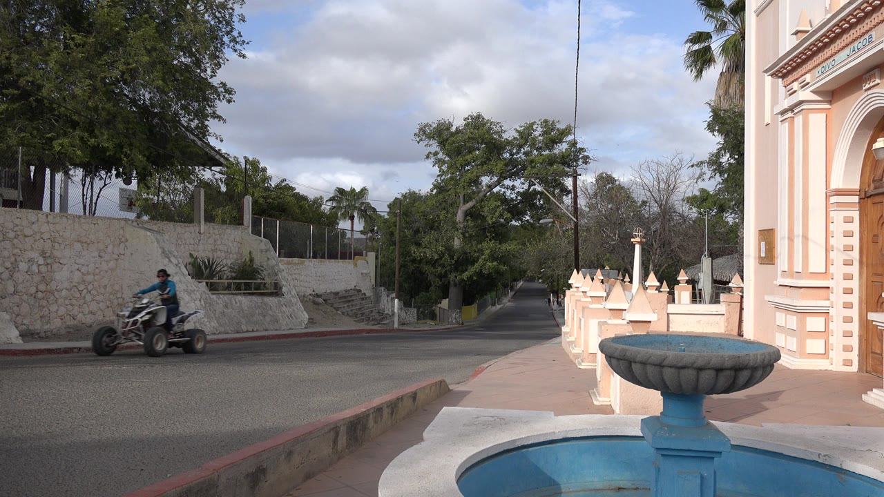 The quad rider continues across the left side of the Los Cabos street, while the blue fountain, building edge, stone retaining wall, trees, and overcast sky remain in view.