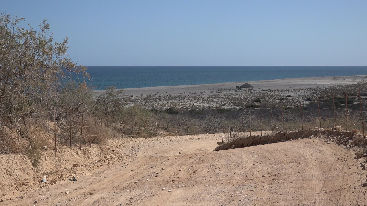Opening frame in Cabo Pulmo, Mexico showing a dusty road, dry brush, wire fencing, rocky beach flats, blue ocean, and no visible people.