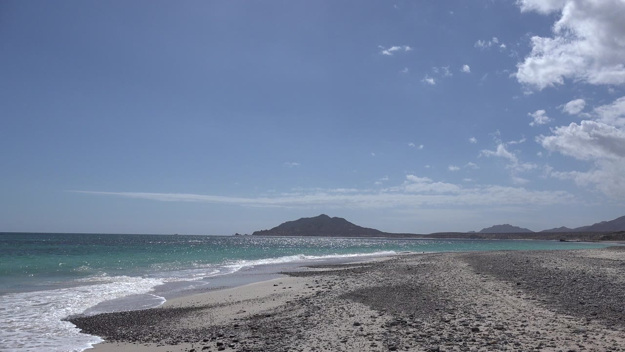 At the start of the Cabo Pulmo clip, white surf reaches the rocky sand with turquoise water and low hills beyond, with no visible people.
