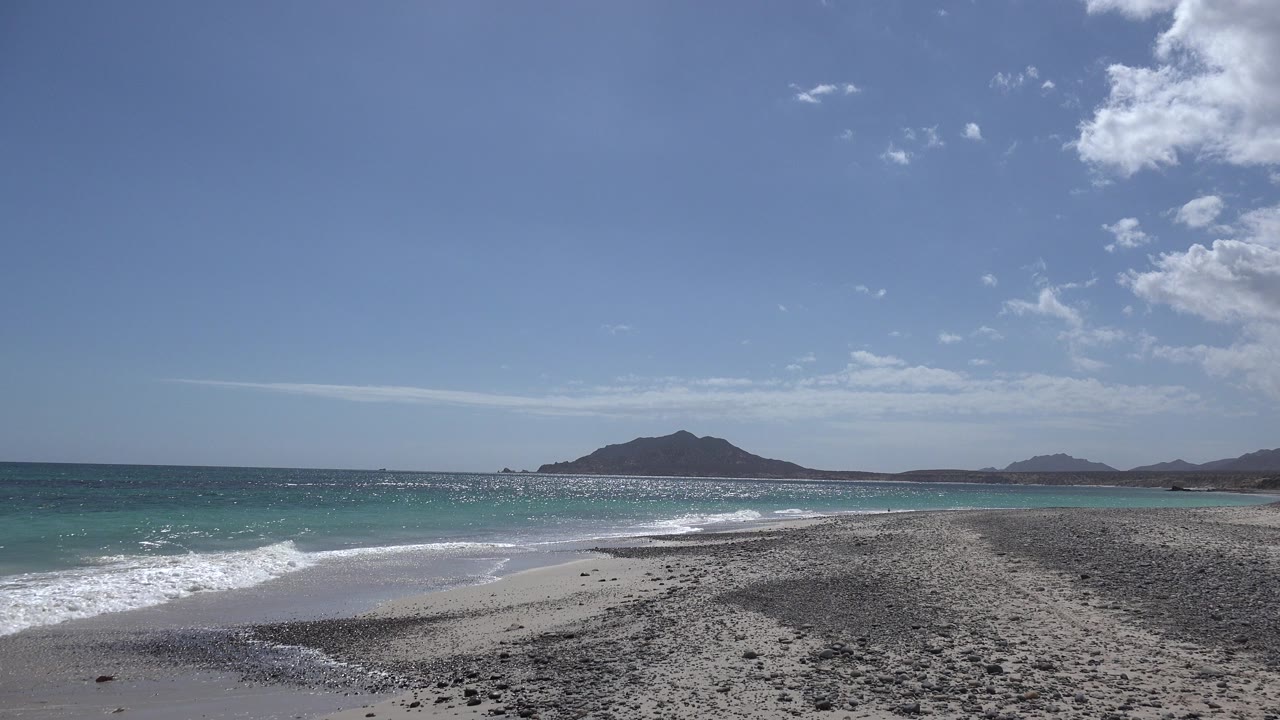 Cabo Pulmo beach at the start of the clip, with dark pebbly sand in the foreground, surf on the left, distant low hills on the horizon, and no visible people.