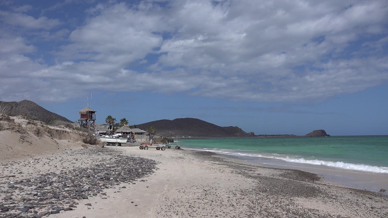 Cabo Pulmo beach at the start of the clip, with pebbles across the foreground sand, turquoise water to the right, low hills beyond the shore, a small raised beach structure near palms, and no visible people.